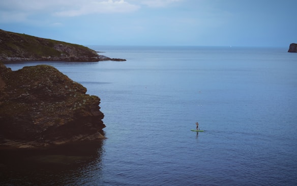 A lone paddleboarder glides across a calm, expansive body of water, surrounded by rocky cliffs on either side. The scene is tranquil, with a distant horizon under a cloudy sky.