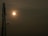 A telecommunications tower stands silhouetted against the sky with a bright sun casting a warm glow. The sky is slightly overcast, adding a soft haze around the sun.
