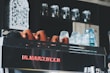 A coffee machine topped with a row of upside-down ceramic cups and glass mugs. The machine is branded with the name 'La Marzocco' in red letters. In the background, there are shelves holding multiple glass jars, adding to a cozy café ambiance.