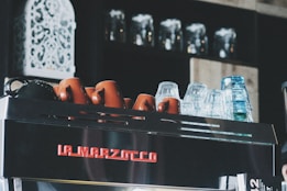 A coffee machine topped with a row of upside-down ceramic cups and glass mugs. The machine is branded with the name 'La Marzocco' in red letters. In the background, there are shelves holding multiple glass jars, adding to a cozy café ambiance.