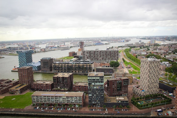 Aerial view of an urban landscape featuring water channels, modern apartment buildings with geometric designs, and surrounding industrial structures. The area is characterized by diverse architectural styles and a mix of green spaces, roads, and residential blocks.