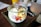 A glass jar filled with yogurt, topped with an assortment of fresh fruits including banana slices, mango cubes, and berries. The jar is placed on a dark tray on a wooden table, with a yellow drink container and a tall glass with a straw visible in the background.