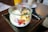 A glass jar filled with yogurt, topped with an assortment of fresh fruits including banana slices, mango cubes, and berries. The jar is placed on a dark tray on a wooden table, with a yellow drink container and a tall glass with a straw visible in the background.