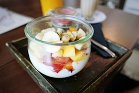 A glass jar filled with yogurt, topped with an assortment of fresh fruits including banana slices, mango cubes, and berries. The jar is placed on a dark tray on a wooden table, with a yellow drink container and a tall glass with a straw visible in the background.
