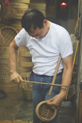 A person is working with a bamboo tool, appearing focused and engaged in the task. The background shows stacks of bamboo baskets, suggesting a workshop or crafting environment. The individual is wearing a white t-shirt and jeans, indicating a casual setting.