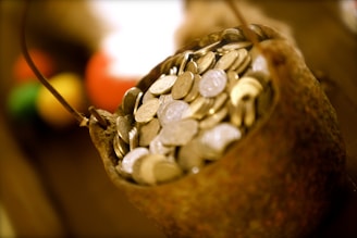A rustic container filled to the brim with numerous coins, suggesting a sense of abundance and wealth. The coins appear metallic, and the container has a rough, textured surface. The background is blurred, but hints of various warm colors can be seen, adding depth to the image.