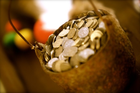 A rustic container filled to the brim with numerous coins, suggesting a sense of abundance and wealth. The coins appear metallic, and the container has a rough, textured surface. The background is blurred, but hints of various warm colors can be seen, adding depth to the image.