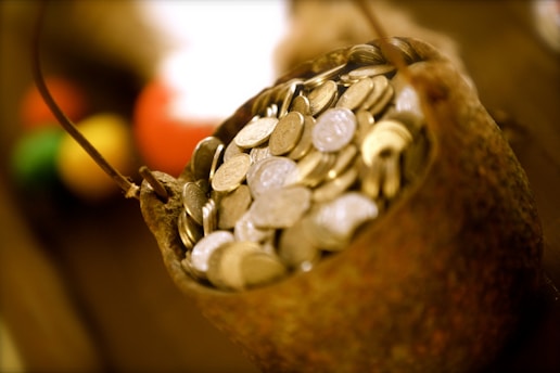 A rustic container filled to the brim with numerous coins, suggesting a sense of abundance and wealth. The coins appear metallic, and the container has a rough, textured surface. The background is blurred, but hints of various warm colors can be seen, adding depth to the image.