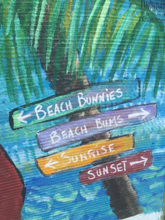 A colorful painted signpost indicating various destinations, including Beach Bunnies, Beach Bums, Sunrise, and Sunset. The signs are attached to a palm tree with vibrant brushstrokes representing green palm leaves in the background, and a blue sky with swirling patterns of white and turquoise suggesting the ocean.