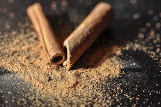 Two cinnamon sticks are placed on a dark surface with scattered cinnamon powder around them. The lighting creates a warm and inviting atmosphere, highlighting the texture of the sticks and the fine granules of the powder.