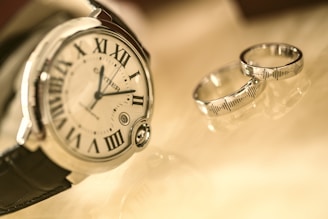 A close-up of a luxury wristwatch with a leather strap, displaying roman numerals on its face. Alongside the watch are two silver rings, positioned on a glossy surface that reflects a warm, golden light.