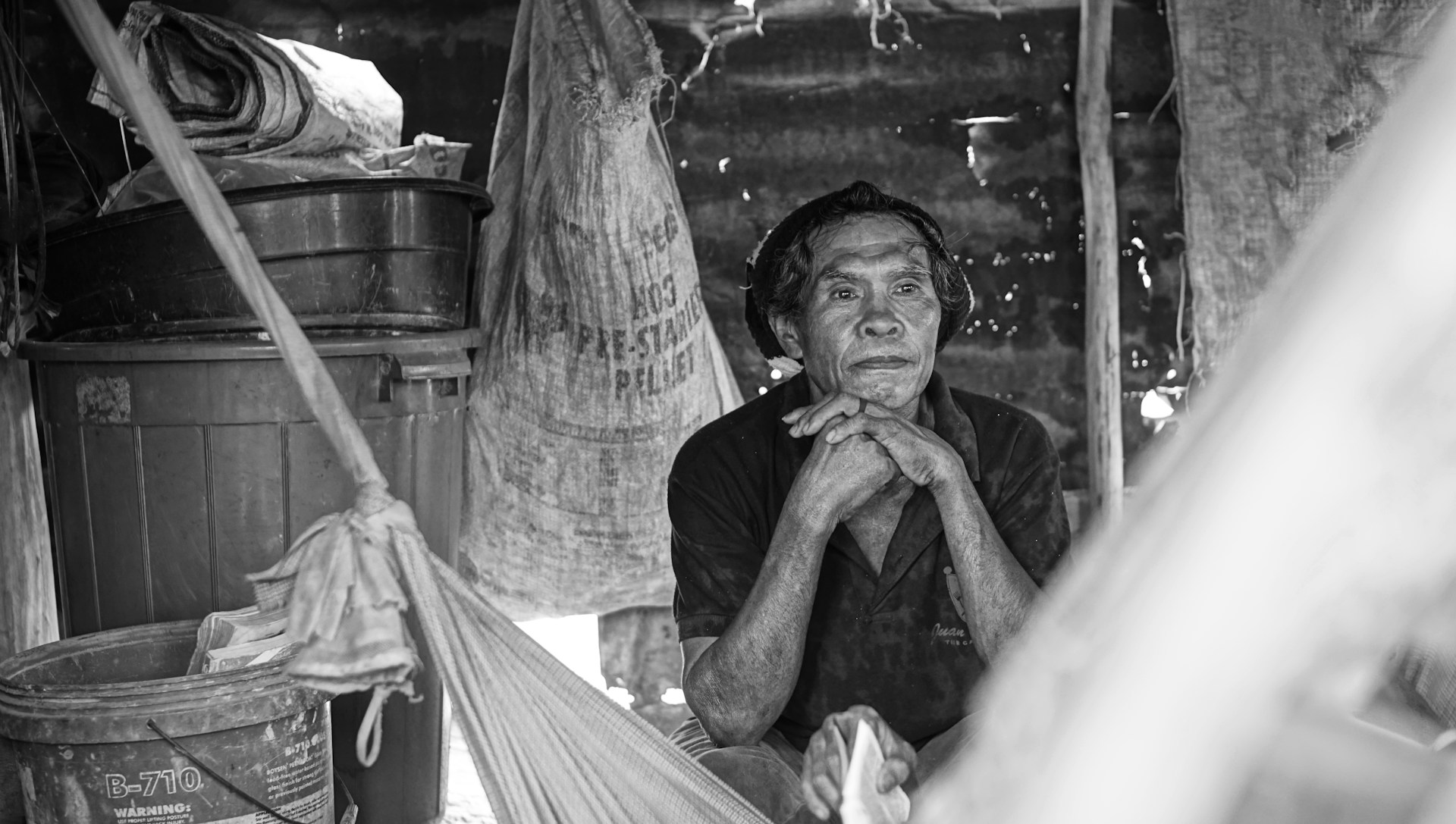 A man sits contemplatively inside a rustic, makeshift shelter made of various materials. His chin rests on his hands, and his expression suggests deep thought or reflection. Surrounding him are items such as large containers, bags, and other household objects.
