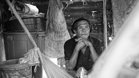 A man sits contemplatively inside a rustic, makeshift shelter made of various materials. His chin rests on his hands, and his expression suggests deep thought or reflection. Surrounding him are items such as large containers, bags, and other household objects.