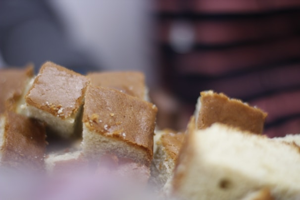 A close-up image of several pieces of light brown cake, cut into square portions. The texture appears soft and spongy, with a slight sheen on the surface suggesting freshness.