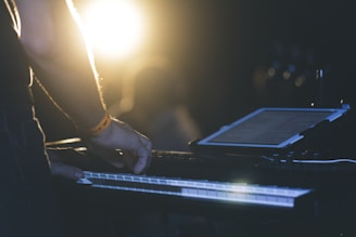 A person is standing in a dimly lit environment, playing a keyboard. A tablet displaying text is positioned on the keyboard stand, and there is a bright light in the background, creating a dramatic silhouette effect.