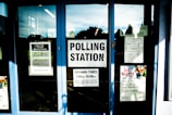 Glass doors with multiple signs indicating polling station information, operating hours, and notices. Various posters are visible with text and images, including one about a local event. The space behind the glass appears to be dimly lit.
