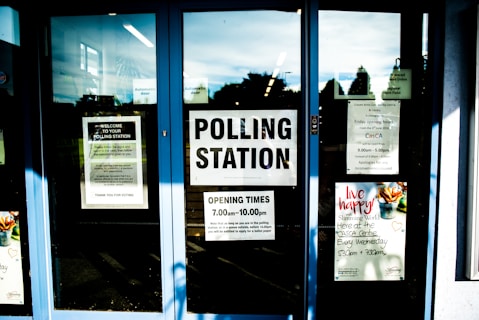 Glass doors with multiple signs indicating polling station information, operating hours, and notices. Various posters are visible with text and images, including one about a local event. The space behind the glass appears to be dimly lit.