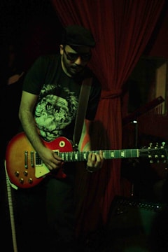 A musician wearing sunglasses and a hat is playing an electric guitar. The setting appears to be dimly lit, with red curtains in the background suggesting a stage or performance area. The musician is focused on playing, and an amplifier is partially visible in the lower part of the image.