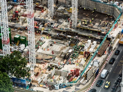 A large construction site featuring multiple high cranes, with numerous workers wearing high-visibility clothing engaged in various tasks. The site has a mix of building materials, machinery, and construction equipment scattered throughout. Adjacent to the site is a busy road with several vehicles, including cars and trucks.