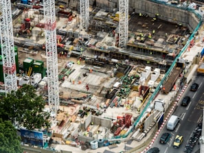 A large construction site featuring multiple high cranes, with numerous workers wearing high-visibility clothing engaged in various tasks. The site has a mix of building materials, machinery, and construction equipment scattered throughout. Adjacent to the site is a busy road with several vehicles, including cars and trucks.