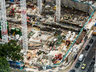 A large construction site featuring multiple high cranes, with numerous workers wearing high-visibility clothing engaged in various tasks. The site has a mix of building materials, machinery, and construction equipment scattered throughout. Adjacent to the site is a busy road with several vehicles, including cars and trucks.