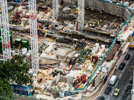A large construction site featuring multiple high cranes, with numerous workers wearing high-visibility clothing engaged in various tasks. The site has a mix of building materials, machinery, and construction equipment scattered throughout. Adjacent to the site is a busy road with several vehicles, including cars and trucks.