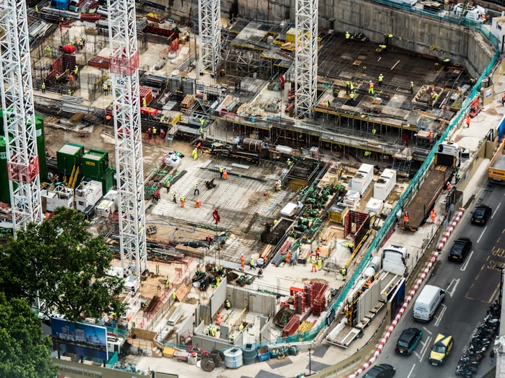A large construction site featuring multiple high cranes, with numerous workers wearing high-visibility clothing engaged in various tasks. The site has a mix of building materials, machinery, and construction equipment scattered throughout. Adjacent to the site is a busy road with several vehicles, including cars and trucks.