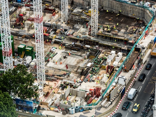 A large construction site featuring multiple high cranes, with numerous workers wearing high-visibility clothing engaged in various tasks. The site has a mix of building materials, machinery, and construction equipment scattered throughout. Adjacent to the site is a busy road with several vehicles, including cars and trucks.