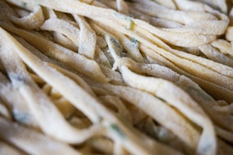 A close-up view of uncooked fettuccine pasta dusted with flour. The pasta strands are light beige with hints of green, likely from added herbs or vegetables, creating a soft, textured appearance.