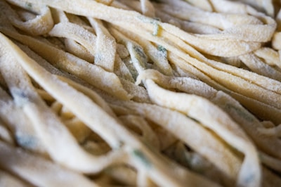 A close-up view of uncooked fettuccine pasta dusted with flour. The pasta strands are light beige with hints of green, likely from added herbs or vegetables, creating a soft, textured appearance.