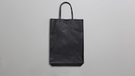 A simple black paper bag with handles is placed against a plain, light gray background. The bag is flat, with minimal texture and shadows, suggesting a straightforward design.