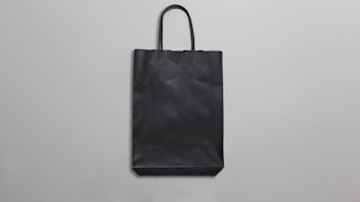 A simple black paper bag with handles is placed against a plain, light gray background. The bag is flat, with minimal texture and shadows, suggesting a straightforward design.