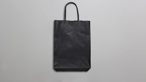 A simple black paper bag with handles is placed against a plain, light gray background. The bag is flat, with minimal texture and shadows, suggesting a straightforward design.