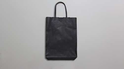 A simple black paper bag with handles is placed against a plain, light gray background. The bag is flat, with minimal texture and shadows, suggesting a straightforward design.