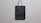 A simple black paper bag with handles is placed against a plain, light gray background. The bag is flat, with minimal texture and shadows, suggesting a straightforward design.