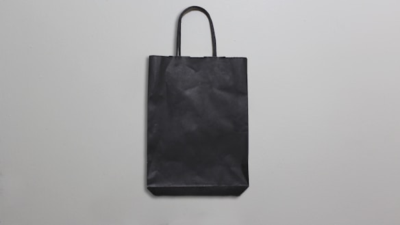 A simple black paper bag with handles is placed against a plain, light gray background. The bag is flat, with minimal texture and shadows, suggesting a straightforward design.