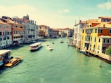 A scenic view of a canal lined with colorful buildings, reflecting the classic architecture of Venice. Several boats, including a water taxi and a larger ferry, navigate the wide, turquoise waters under a clear blue sky.