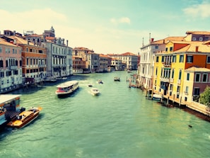 A scenic view of a canal lined with colorful buildings, reflecting the classic architecture of Venice. Several boats, including a water taxi and a larger ferry, navigate the wide, turquoise waters under a clear blue sky.
