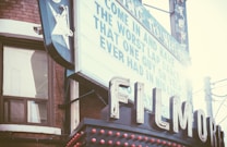 A vintage theater sign with large white letters spelling 'FILMOR'. The marquee above displays a humorous message about a poor experience. The setting includes a brick building with a window and some power lines. Sunlight creates a lens flare effect.