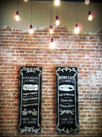 Two vintage-style menus are displayed on a brick wall, featuring decorative chalkboard designs with French text related to a bakery and caf&eacute;. Above, several exposed light bulbs hang from the ceiling, creating a warm and inviting ambiance.