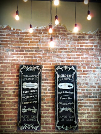 Two vintage-style menus are displayed on a brick wall, featuring decorative chalkboard designs with French text related to a bakery and caf&eacute;. Above, several exposed light bulbs hang from the ceiling, creating a warm and inviting ambiance.