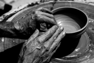 Two hands are skillfully shaping a clay pot on a pottery wheel, displaying the texture of the clay and the hands. The focus is on the intricate details of the potter's hands and the smooth surface of the clay vessel.