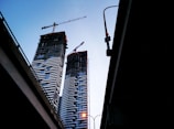 Two high-rise buildings under construction are visible, each equipped with cranes on top. The buildings have a distinct pattern of alternating light and dark horizontal lines. The silhouettes of highway overpasses frame the edges of the image, creating a sense of depth. A streetlamp on one overpass emits a warm glow, contrasting with the early evening sky.