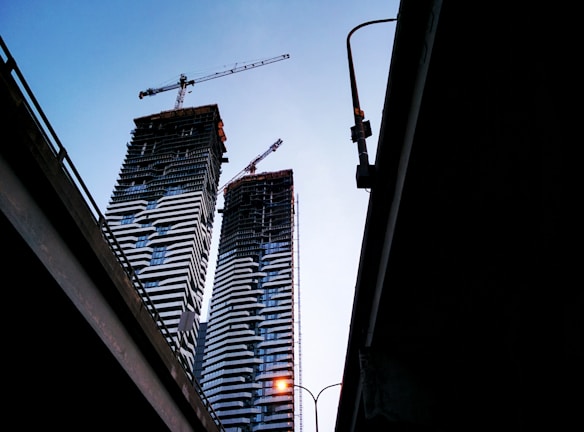 Two high-rise buildings under construction are visible, each equipped with cranes on top. The buildings have a distinct pattern of alternating light and dark horizontal lines. The silhouettes of highway overpasses frame the edges of the image, creating a sense of depth. A streetlamp on one overpass emits a warm glow, contrasting with the early evening sky.