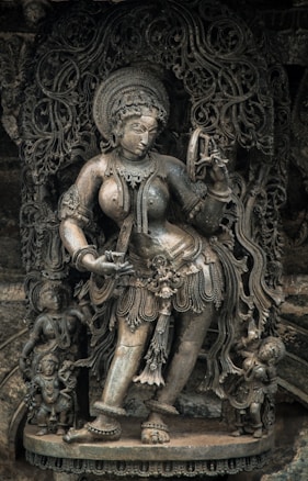 An intricately carved stone sculpture depicts a graceful dancing figure adorned with elaborate jewelry and attire. The figure is surrounded by smaller figures and detailed ornamental patterns, emphasizing traditional artistry.