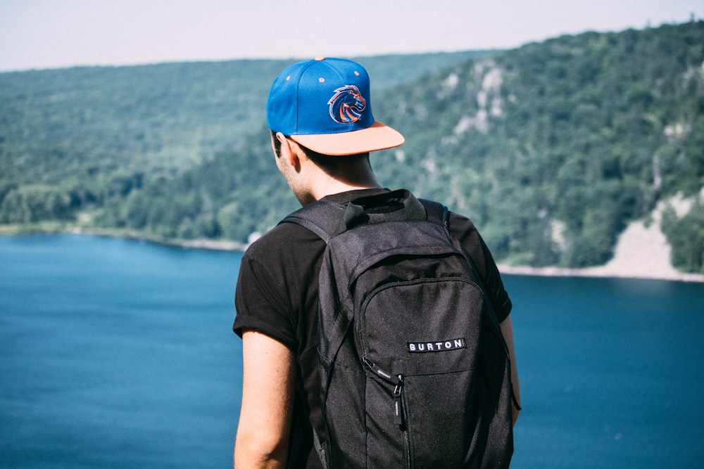 man wearing black t-shirt, black backpack, and blue fitted cap facing body of water photo