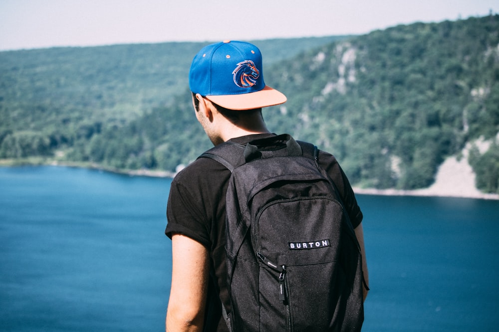 Mann mit schwarzem T-Shirt, schwarzem Rucksack und blauer Mütze mit Blick auf ein Gewässerfoto