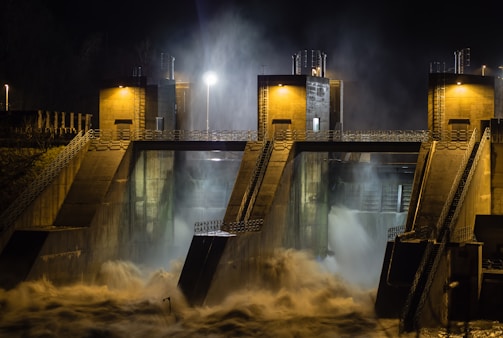 A large dam structure at night, with powerful water flow cascading through its gates. The bright lights illuminate the concrete architecture, creating a dramatic contrast between the dark surroundings and the lit structures. Mist rises from the churning water, adding a sense of movement and intensity.