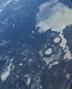 Aerial view of a vast landscape featuring snow-covered areas, bodies of water partially frozen, and dense forested regions. The terrain has a patchwork of dark forest areas contrasting starkly with lighter snow and ice surfaces. The lakes and rivers are interspersed irregularly throughout the scene, creating an intricate pattern within the natural environment.