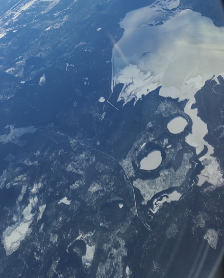 Aerial view of a vast landscape featuring snow-covered areas, bodies of water partially frozen, and dense forested regions. The terrain has a patchwork of dark forest areas contrasting starkly with lighter snow and ice surfaces. The lakes and rivers are interspersed irregularly throughout the scene, creating an intricate pattern within the natural environment.
